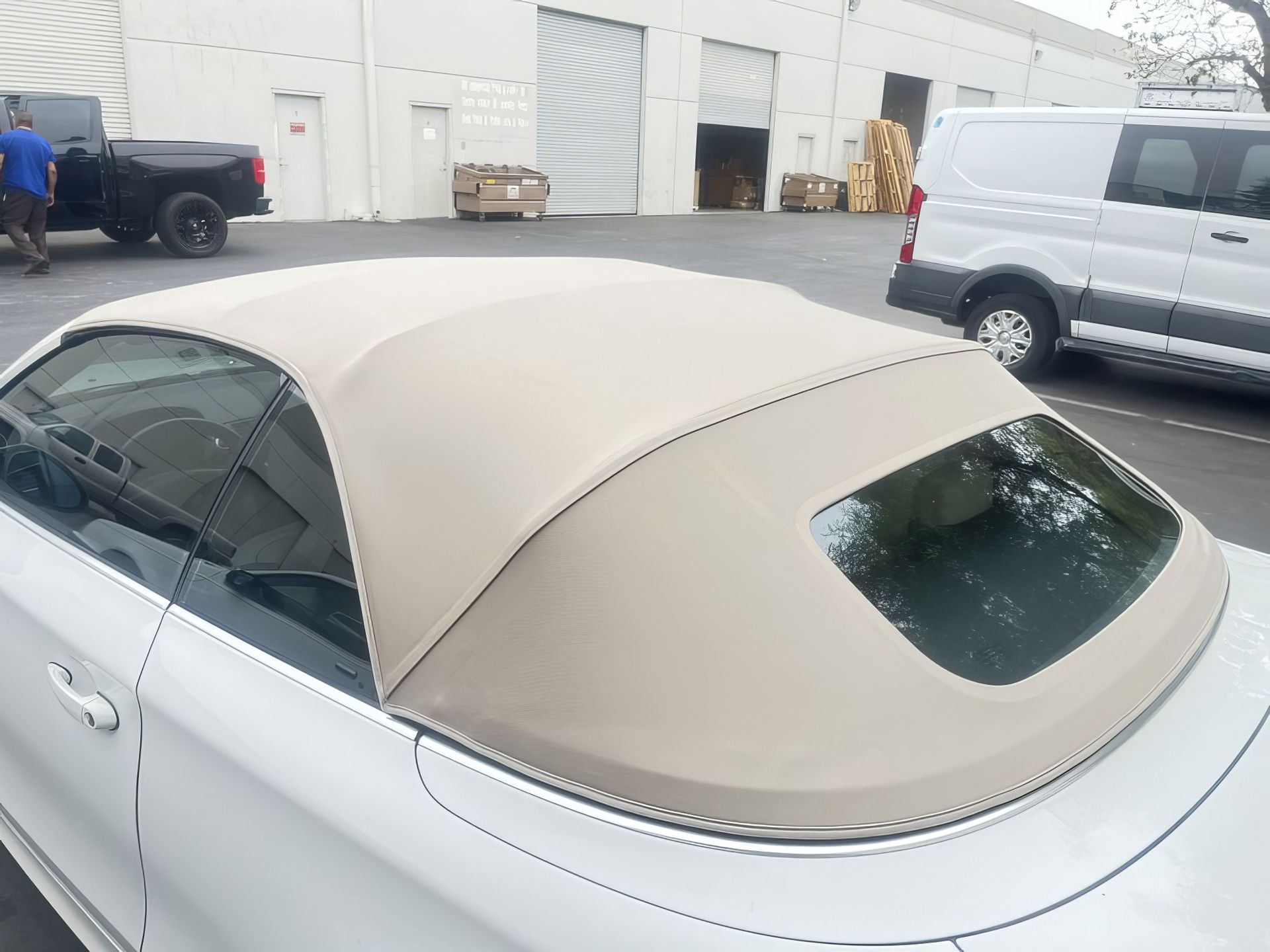 Beige soft-top convertible parked in an industrial lot beside white vans and a blue truck.