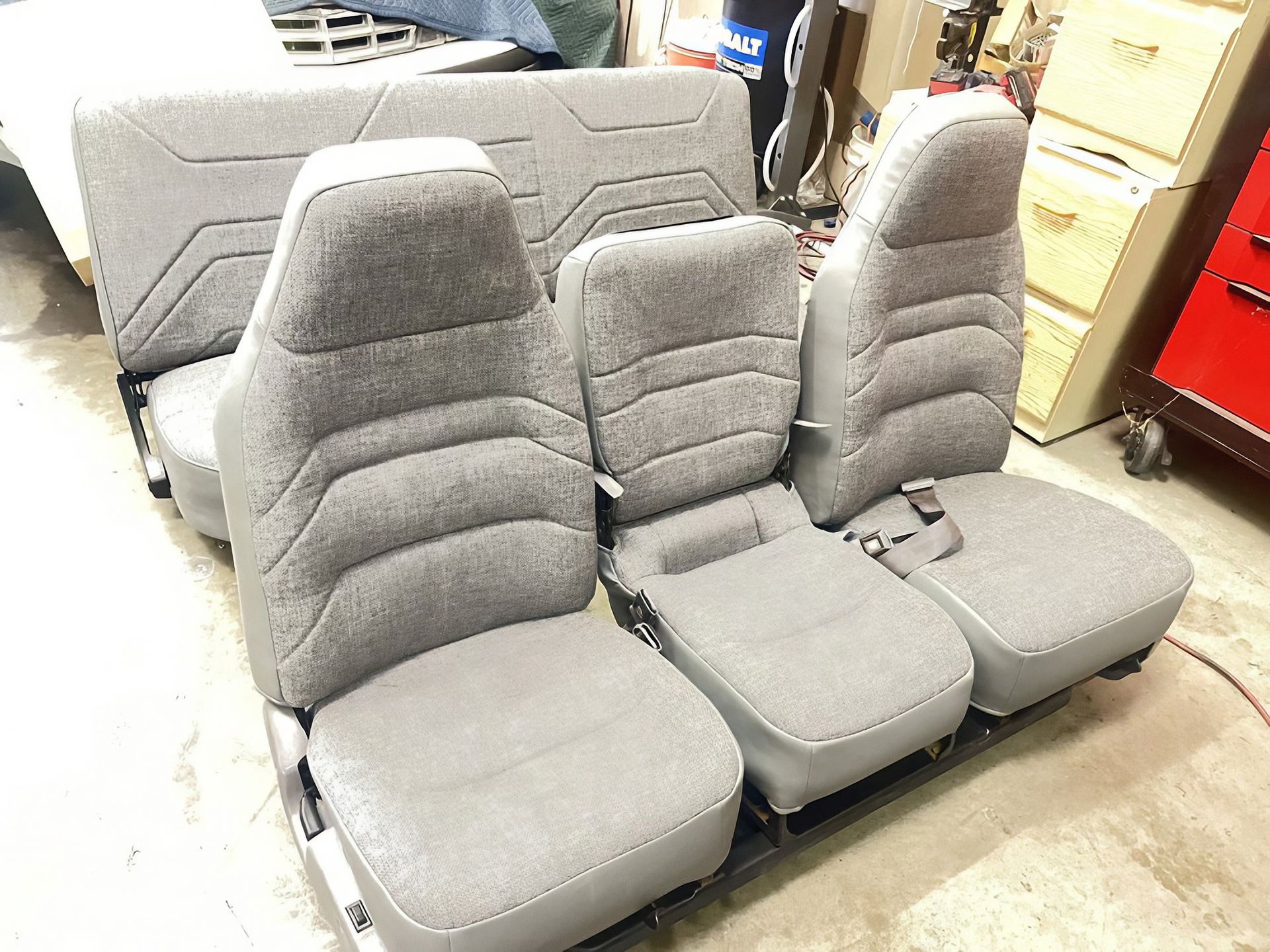 Gray upholstered car seat bench rows in a showroom or workshop, with a couch behind them
