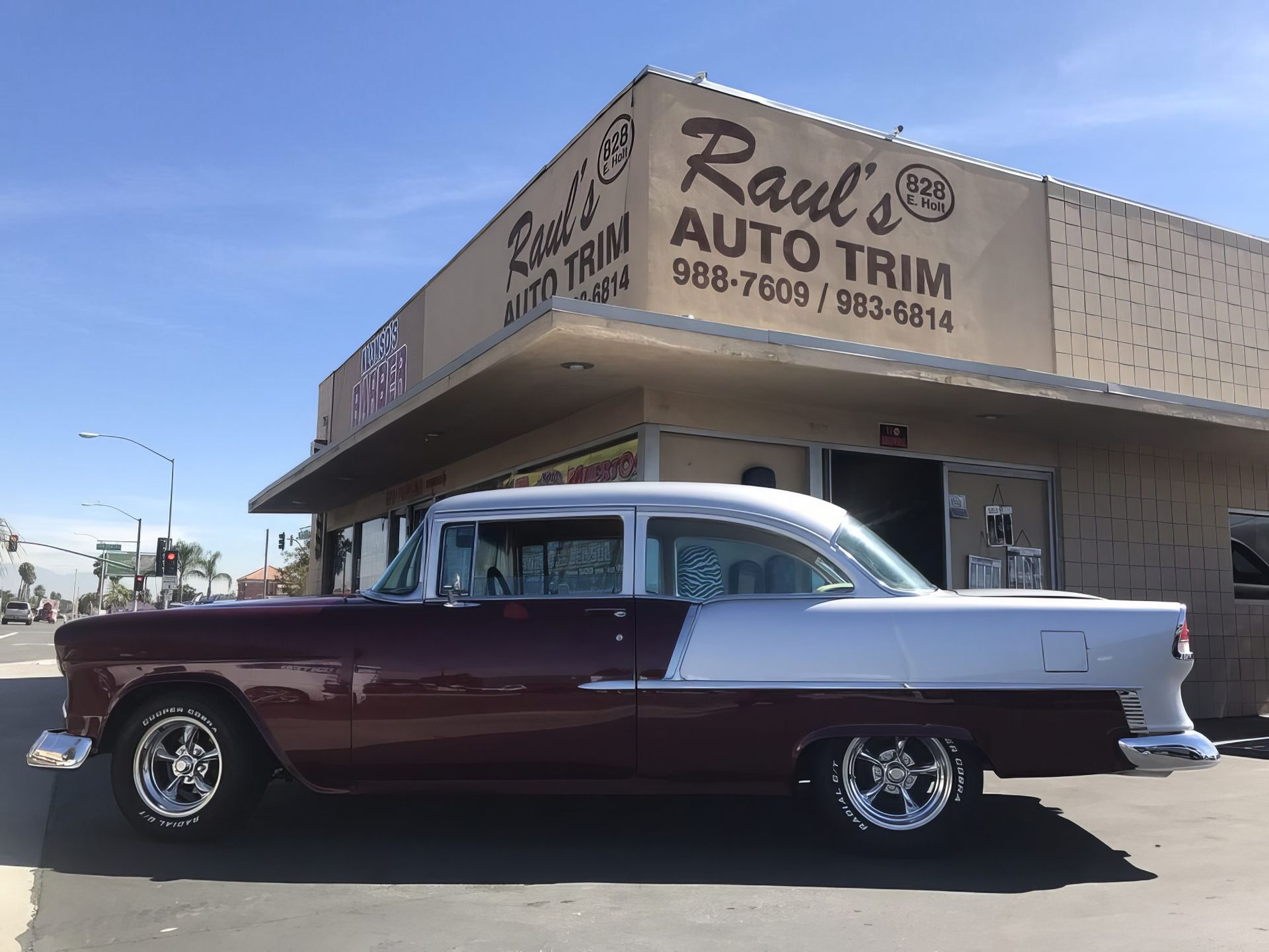 Classic burgundy and white vintage car parked outside a body shop under a blue sky.
