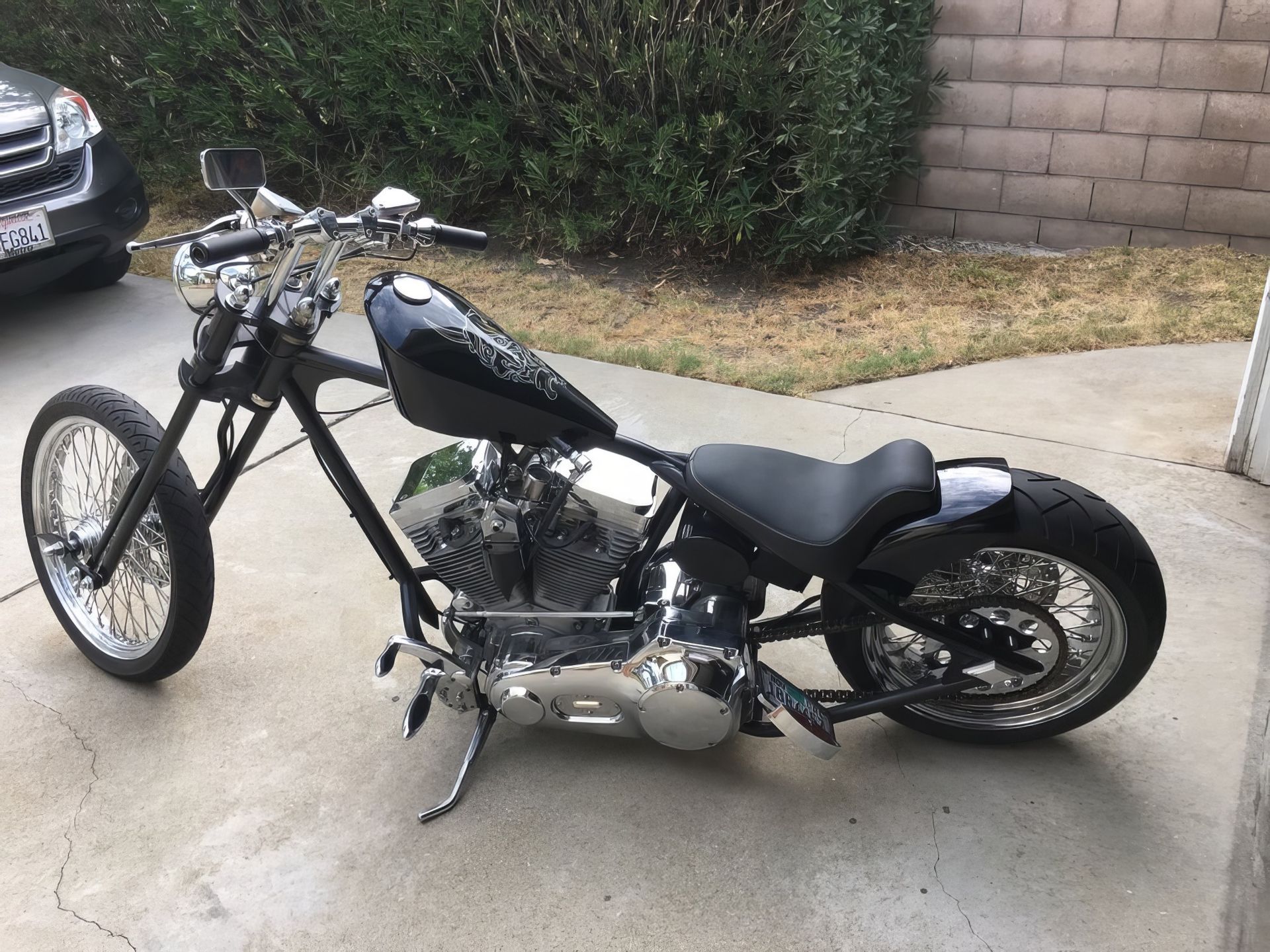 Black custom chopper motorcycle parked on a driveway beside a hedge and brick wall