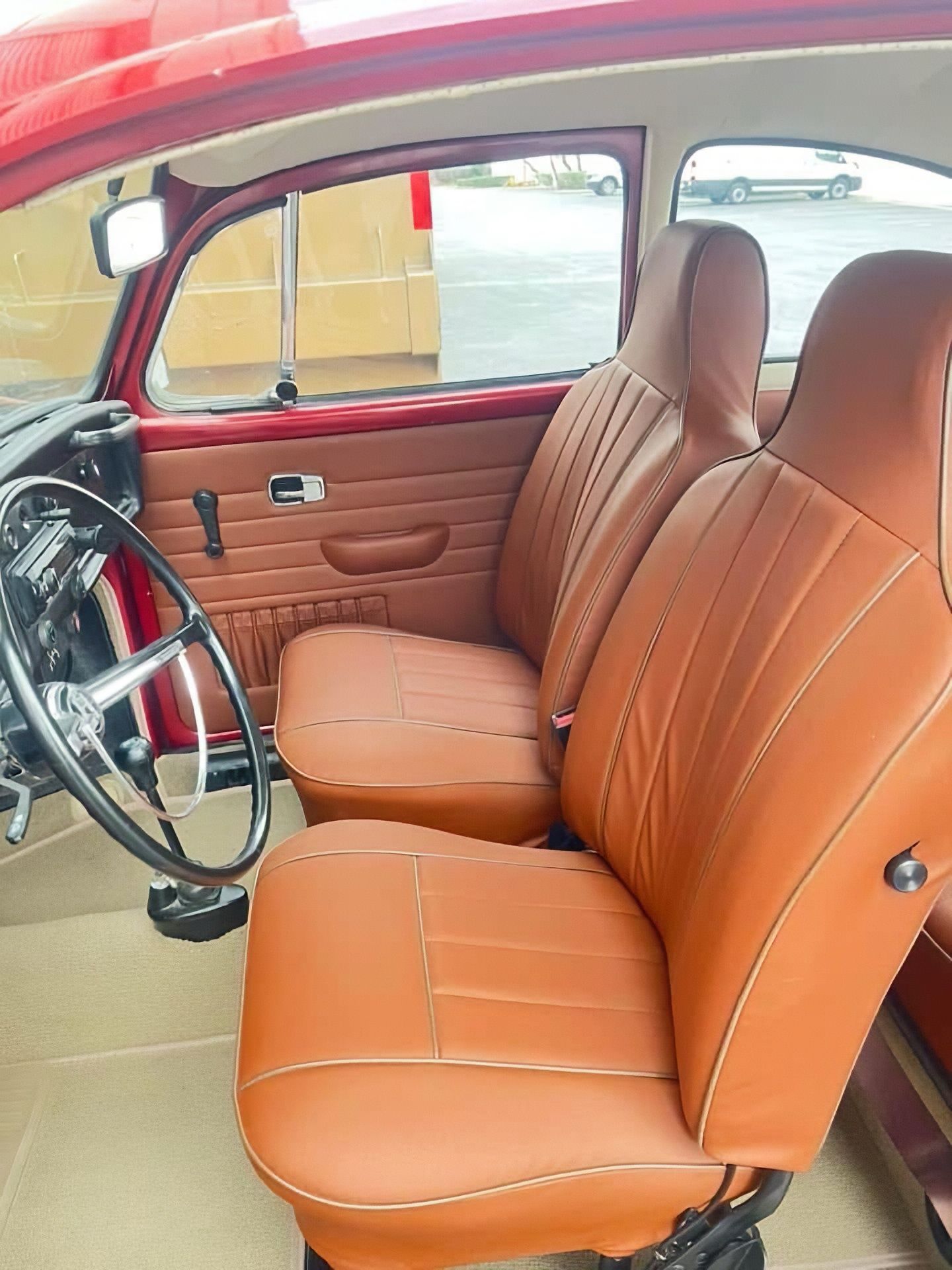 Interior of a vintage car with tan leather seats and a red door panel, viewed from the open driver’s side.