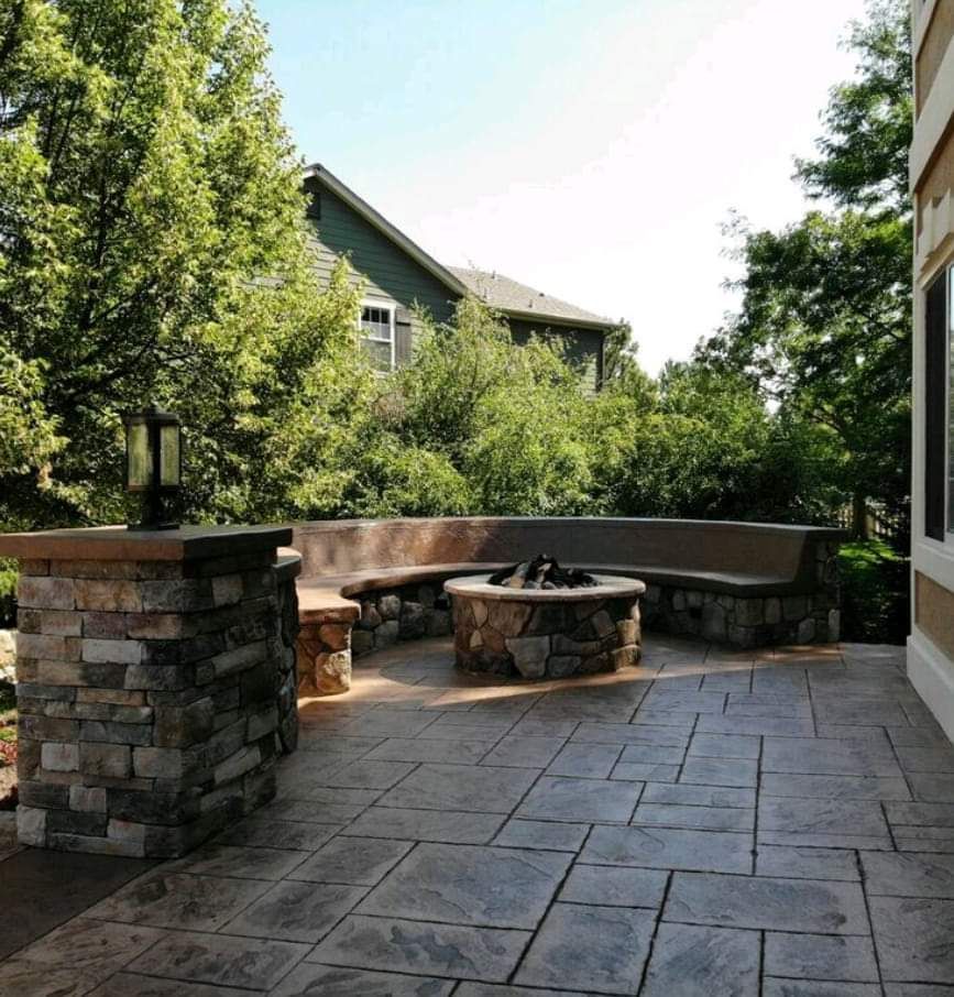 Stone fire pit with built-in bench seating on a stamped concrete patio, surrounded by lush trees and a house.