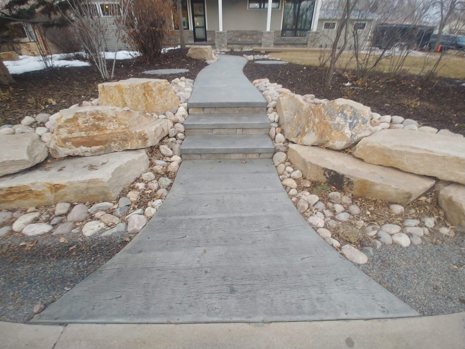 Concrete walkway with steps leading to a house, bordered by large rocks and small pebbles.