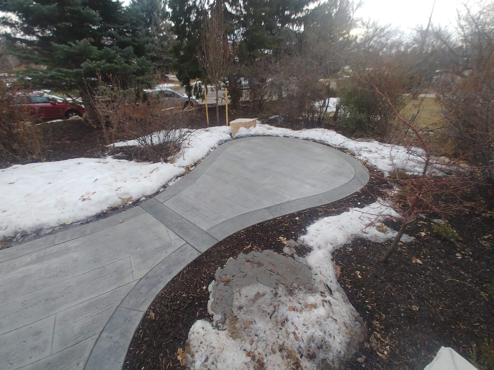 Curving concrete driveway and patio area, edged with dark border and melting snow.