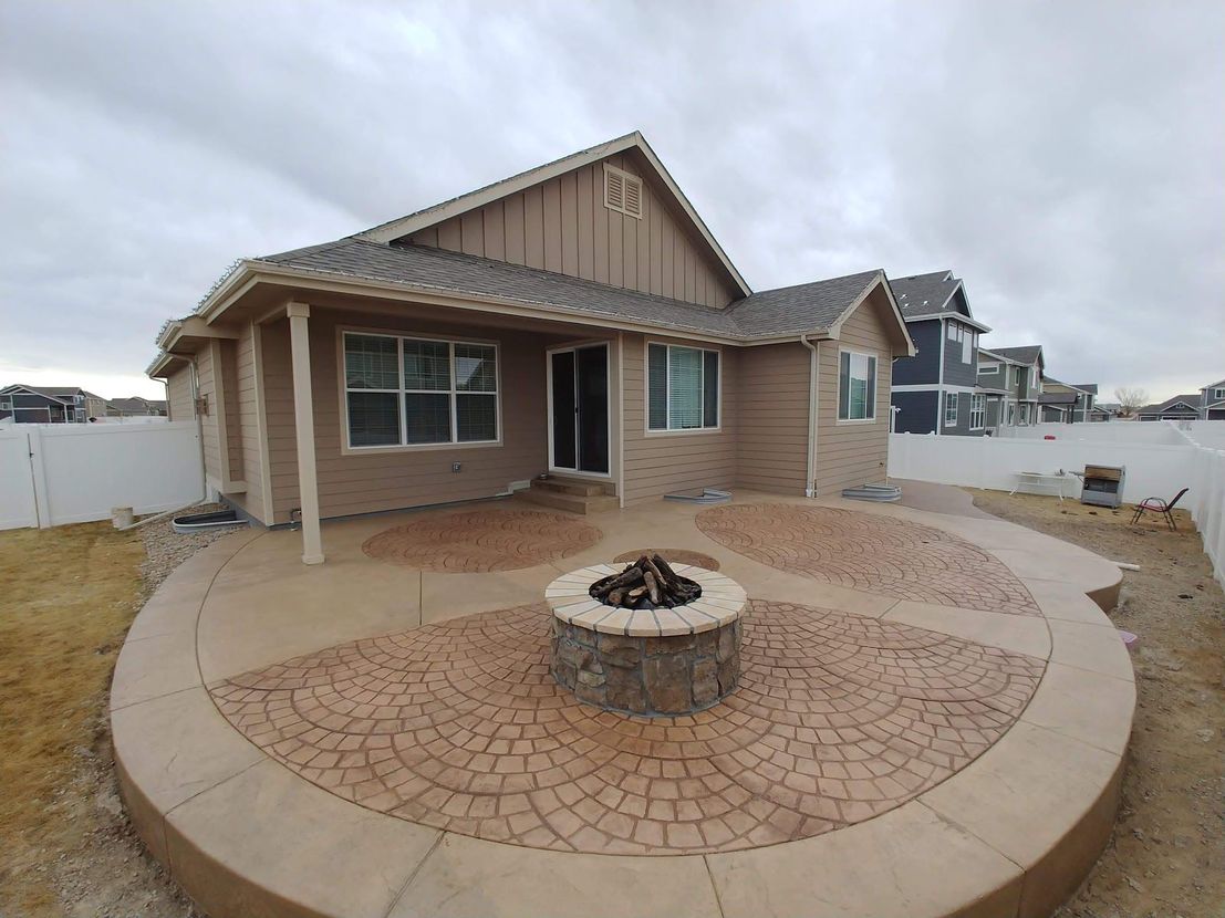 Backyard patio with fire pit, surrounded by patterned concrete and a tan house under a cloudy sky.