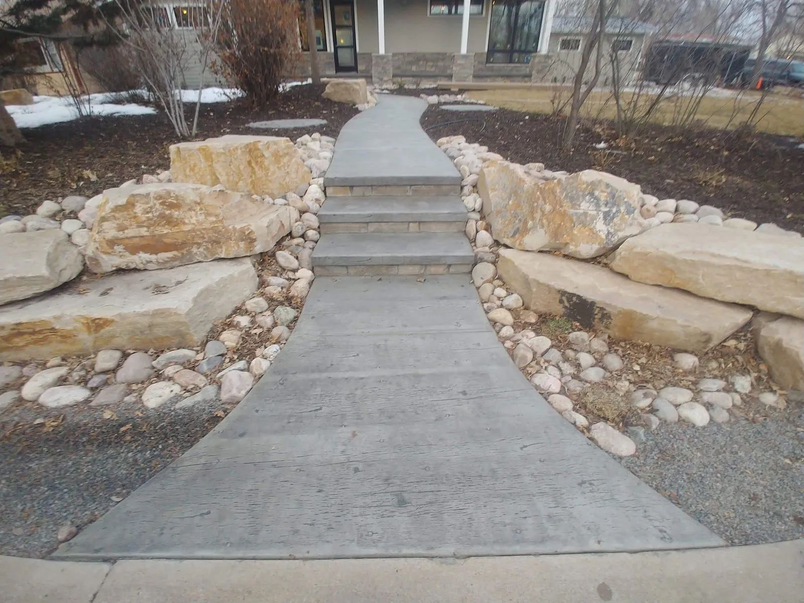 Concrete walkway with steps, lined with large rocks and pebbles, leading to a house.