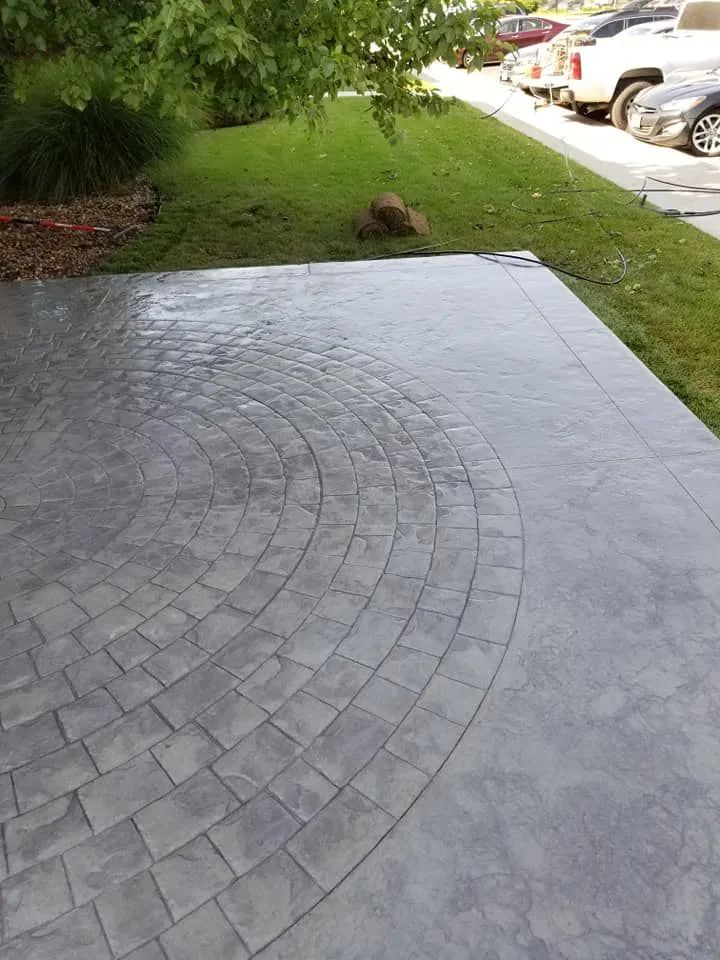 Gray concrete patio with a circular brick pattern, adjacent to a green lawn and sidewalk with parked cars.