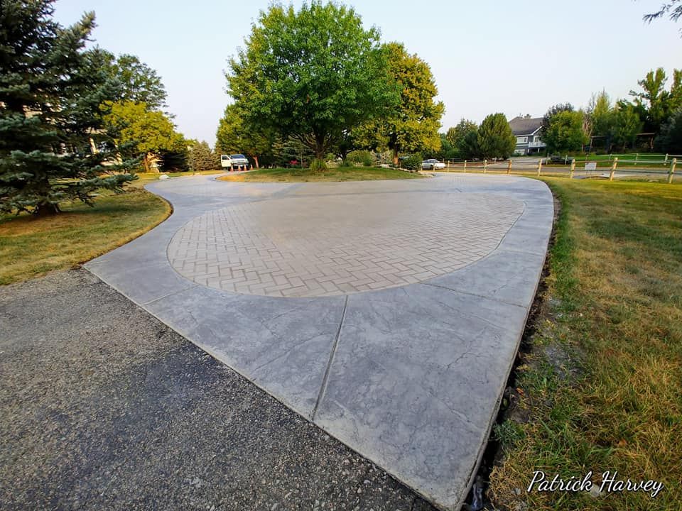 A paved driveway with a circular brick-like pattern, surrounded by grass and trees.