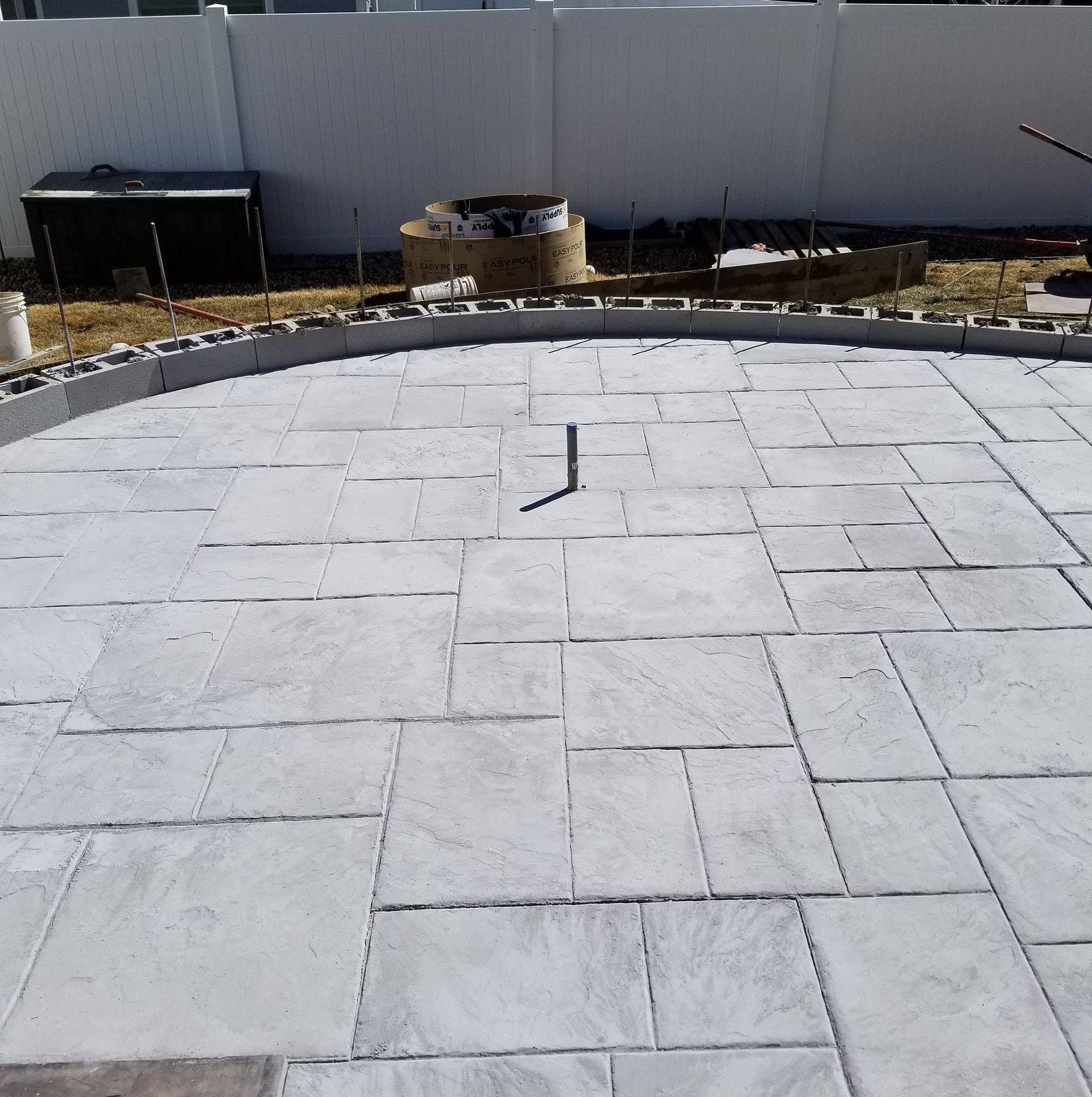 Light gray stone patio with curved edge and a small metal stake in the center, beside a yard and fence.