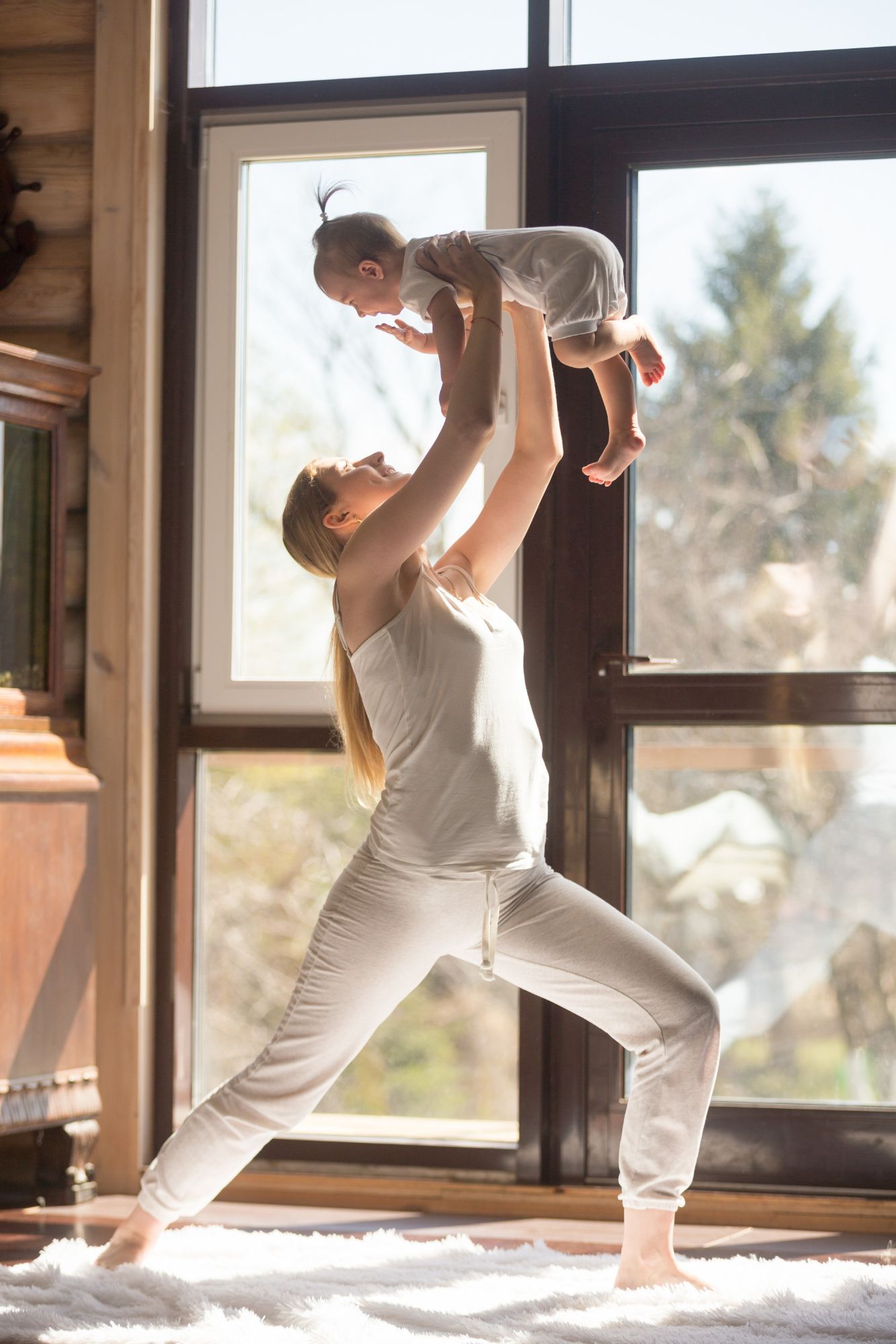 Une femme soulève un bébé devant une fenêtre, pratiquant le yoga dans une pièce lumineuse.