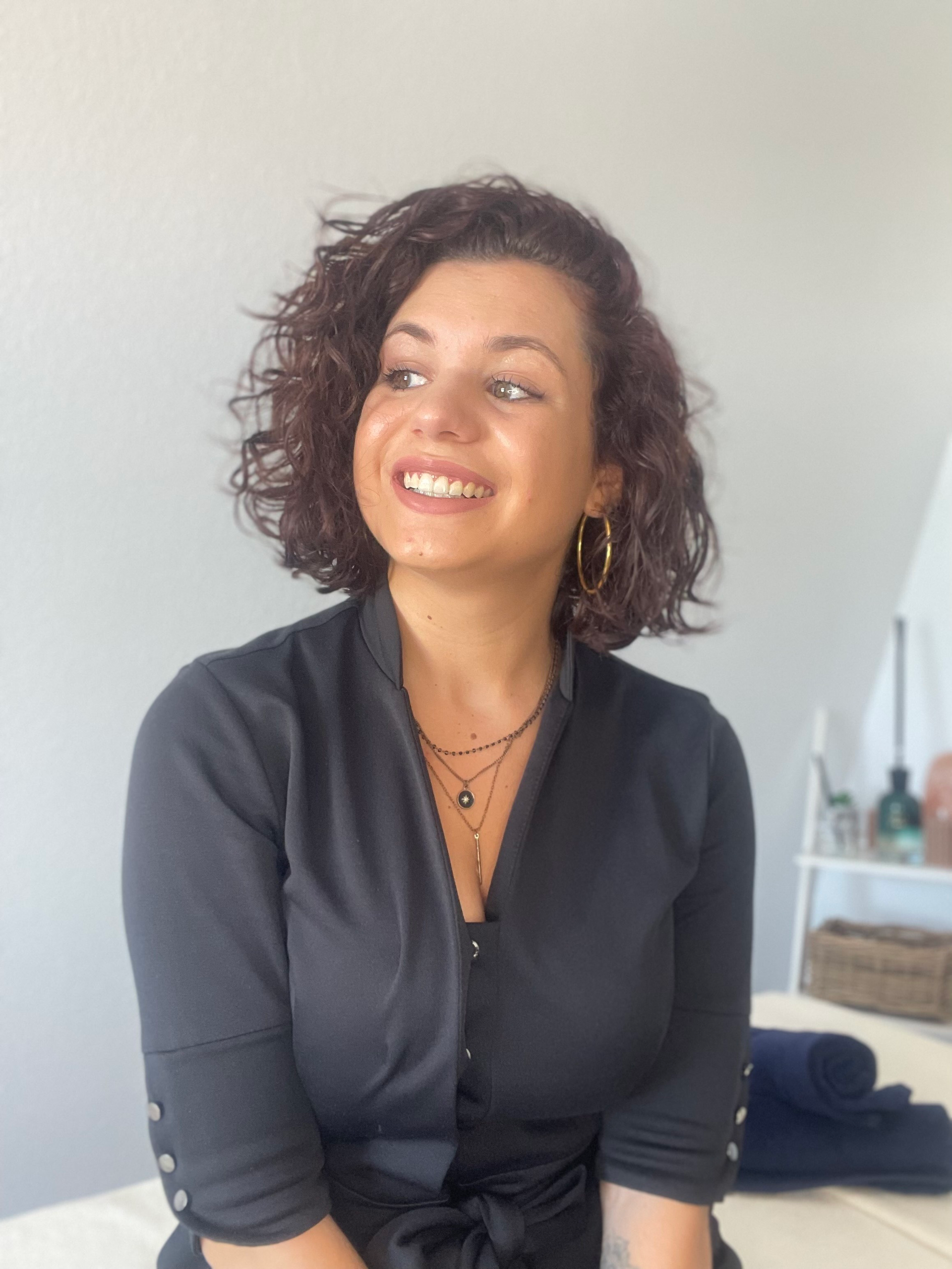Femme aux cheveux bouclés, souriante, vêtue d'un haut noir, assise. Lumière naturelle vive.