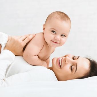 Un bébé sourit, allongé sur une femme ; tous deux sont sur une surface blanche et interagissent.