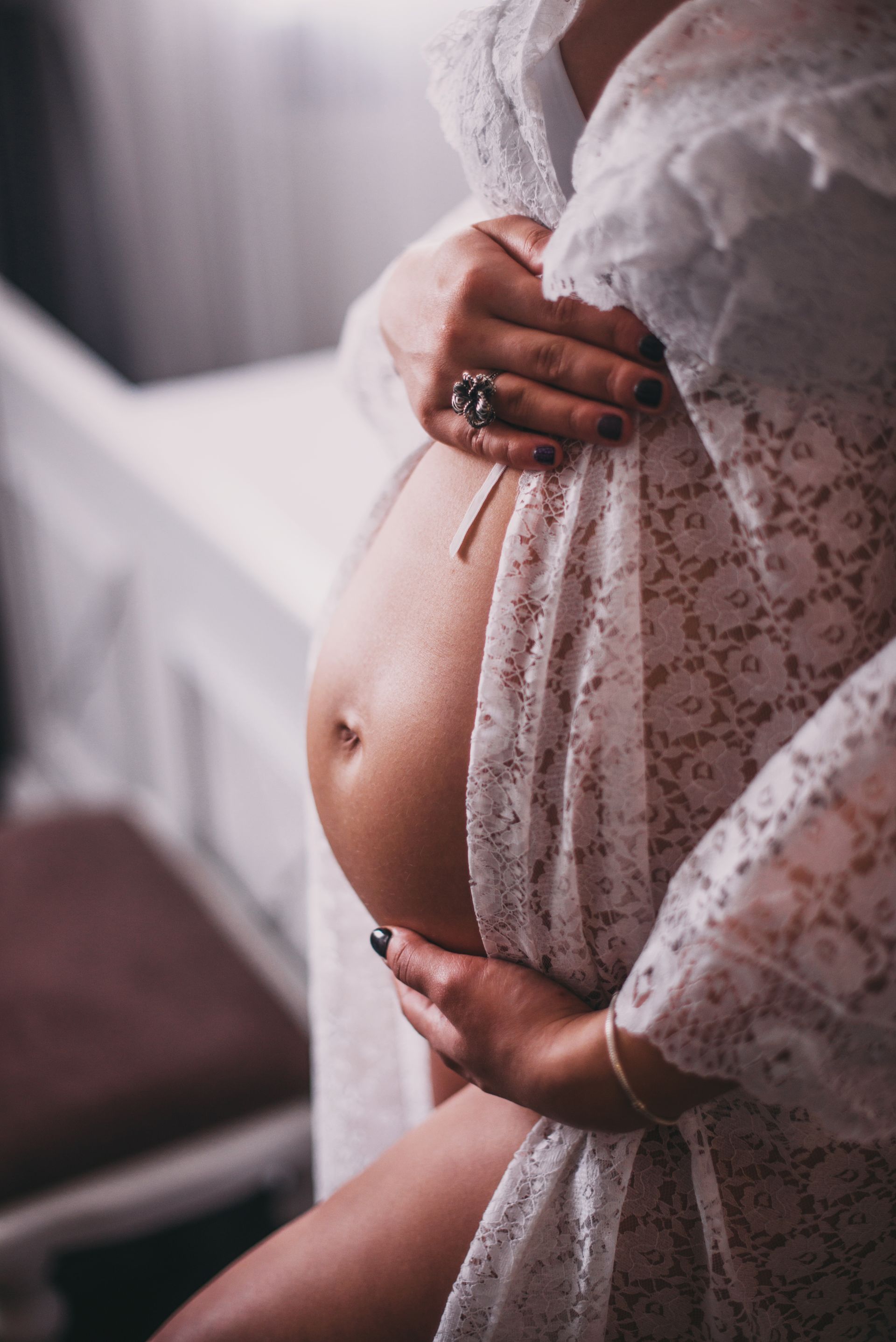 Femme enceinte se tenant le ventre, vêtue d'une robe de dentelle blanche, debout près d'une fenêtre et de meubles.