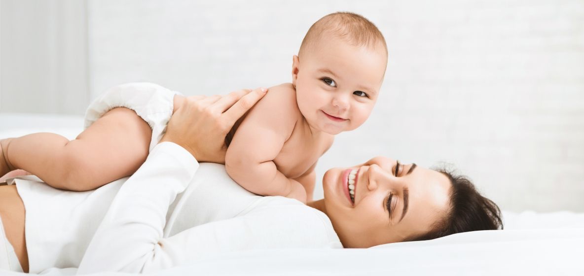 Un bébé rampe sur une femme allongée sur un lit, tous deux souriants.