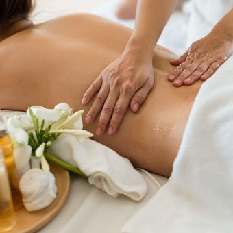 Des mains massent le dos d'une personne, dans une ambiance de spa. Des fleurs et des huiles sont au premier plan.