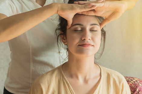 Femme recevant un massage de la tête, les yeux fermés, les mains du thérapeute posées sur son front. Lumière du soleil.
