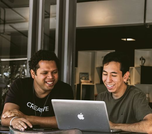 Two men are sitting at a table looking at an apple laptop.