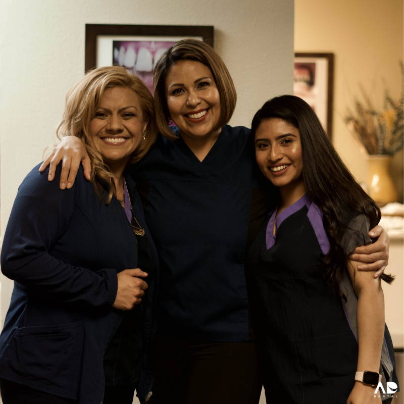 Three women in medical scrubs smile, with arms around each other in a warmly lit office setting.