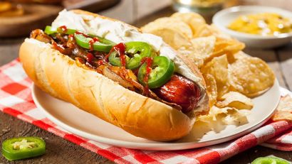 Hot dog with jalapeños and onions on a plate with potato chips, served on a red checkered napkin