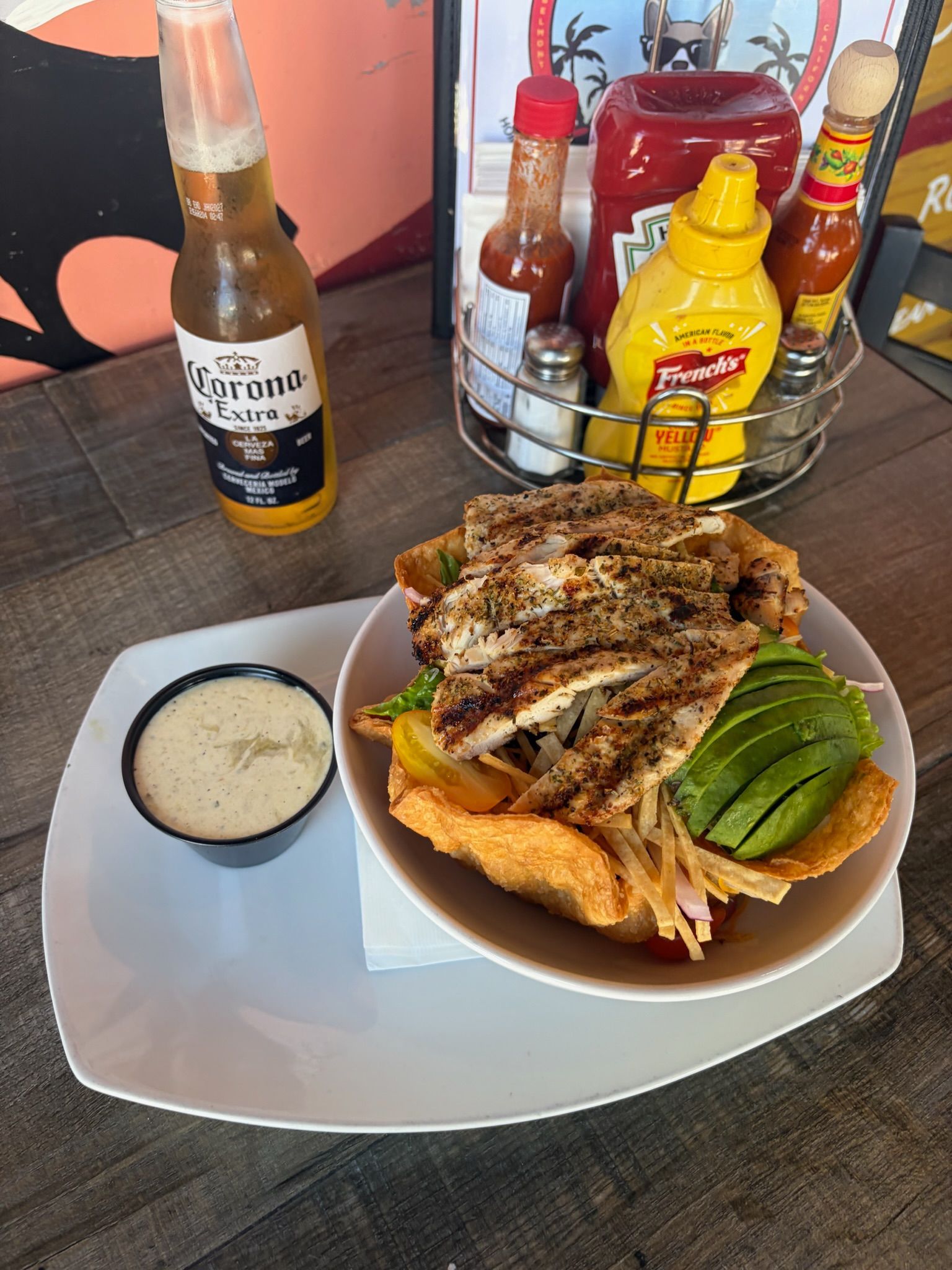 Grilled chicken with tostada, avocado, and sauce on a table beside a Corona beer bottle and condiments.