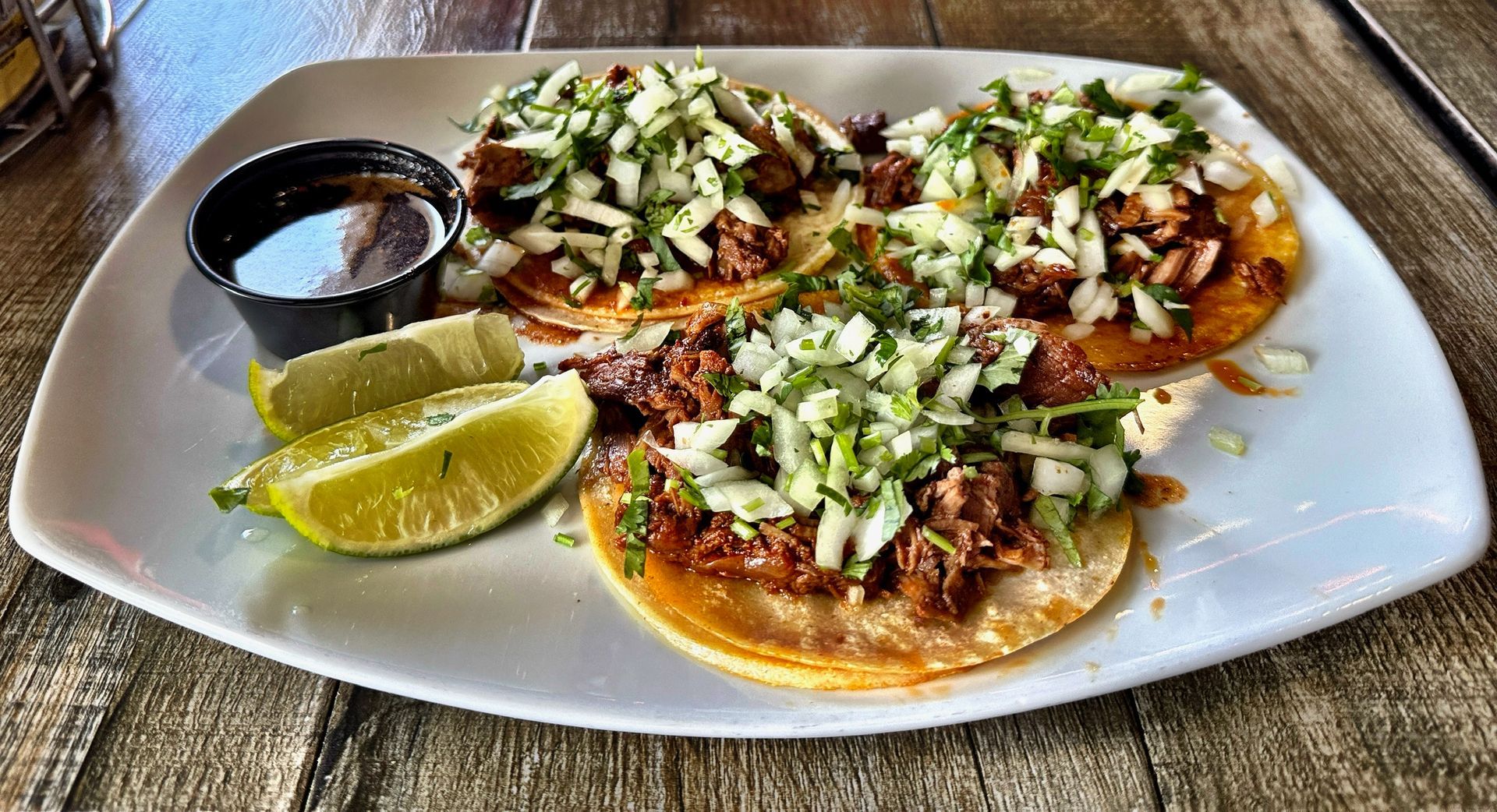 Plate of tostadas with meat, chopped onions, lime wedges, and a small cup of salsa on a wooden table