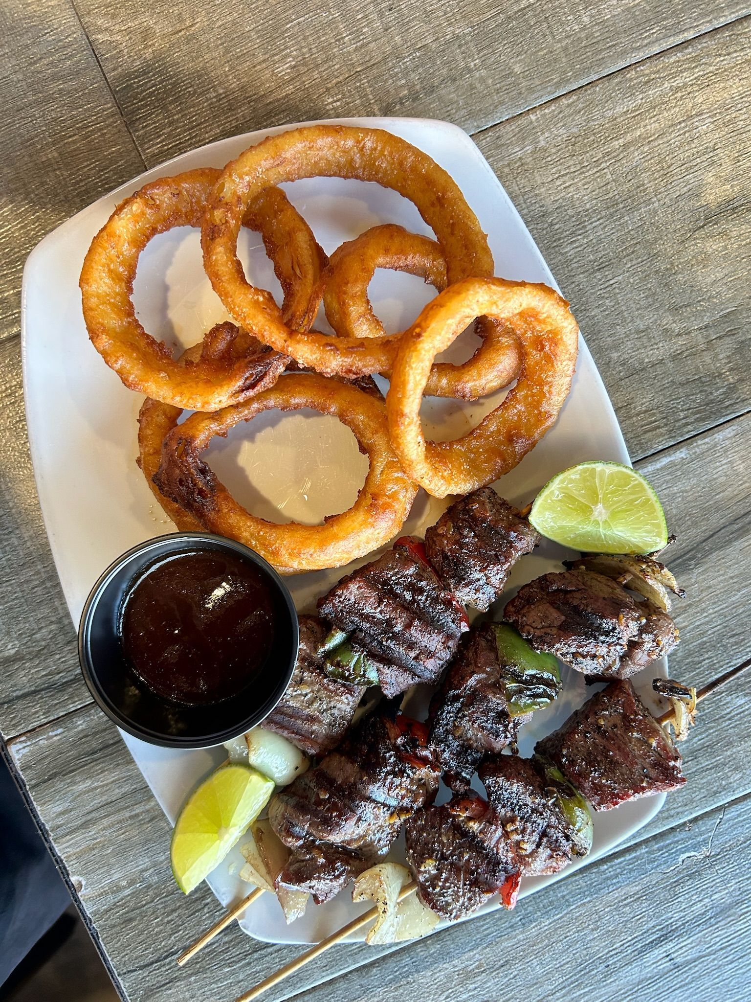 Onion rings and grilled meat skewers with dipping sauce and lime on a white plate.