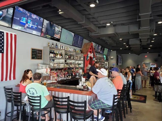 Bar-style restaurant with patrons seated at a counter, TVs overhead, and a large American flag on the wall