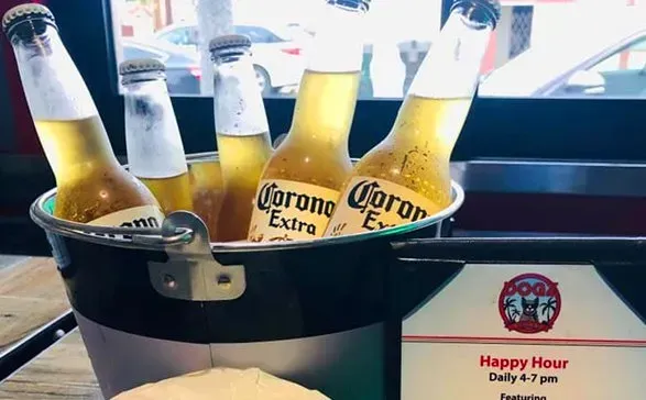 Bucket of Corona beers with lime wedges on a restaurant table, happy hour sign visible in background