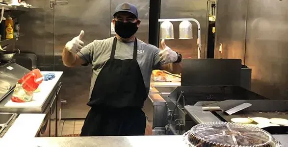 Person in black apron and cap giving thumbs up in a commercial kitchen with food prep and grills.