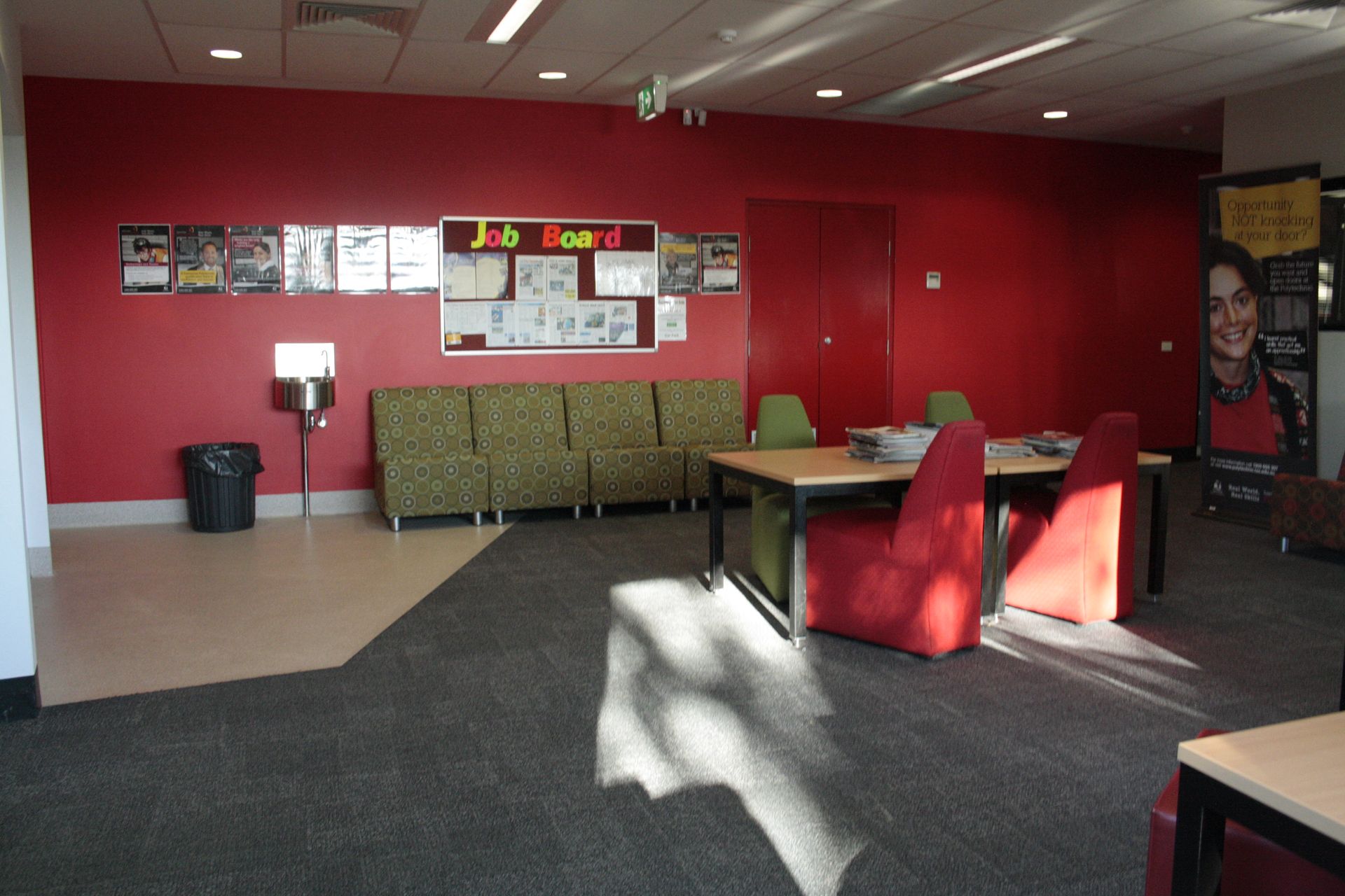Red-walled lobby with chairs, tables, bulletin boards, and sunlight on the carpet — Unique Floors In New Town, TAS