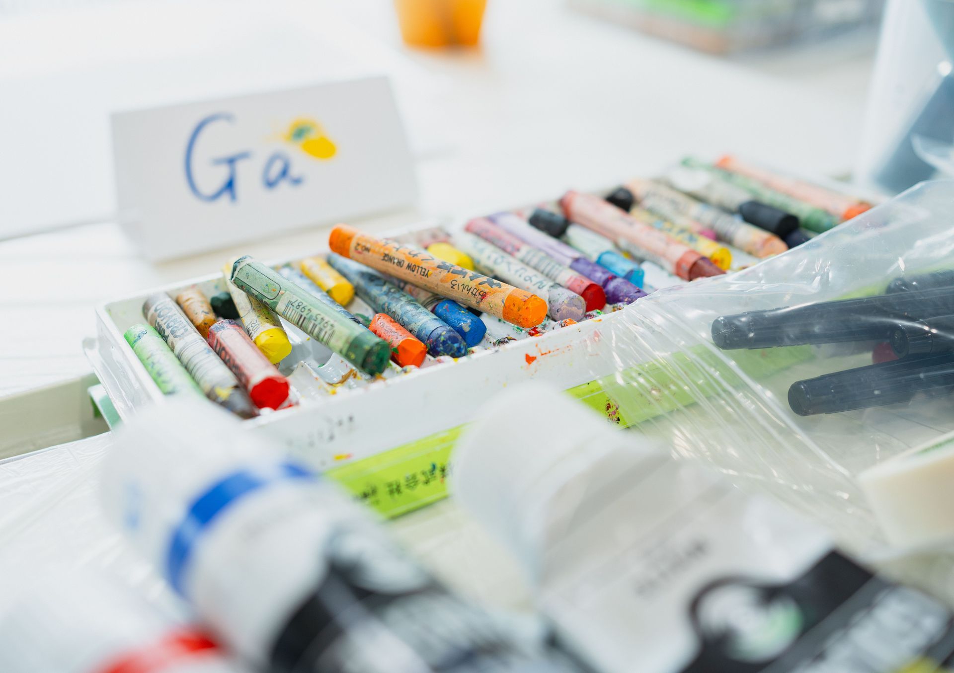 Caja de rotuladores y bolígrafos de colores sobre un escritorio, con una tarjeta con “Ga” al fondo
