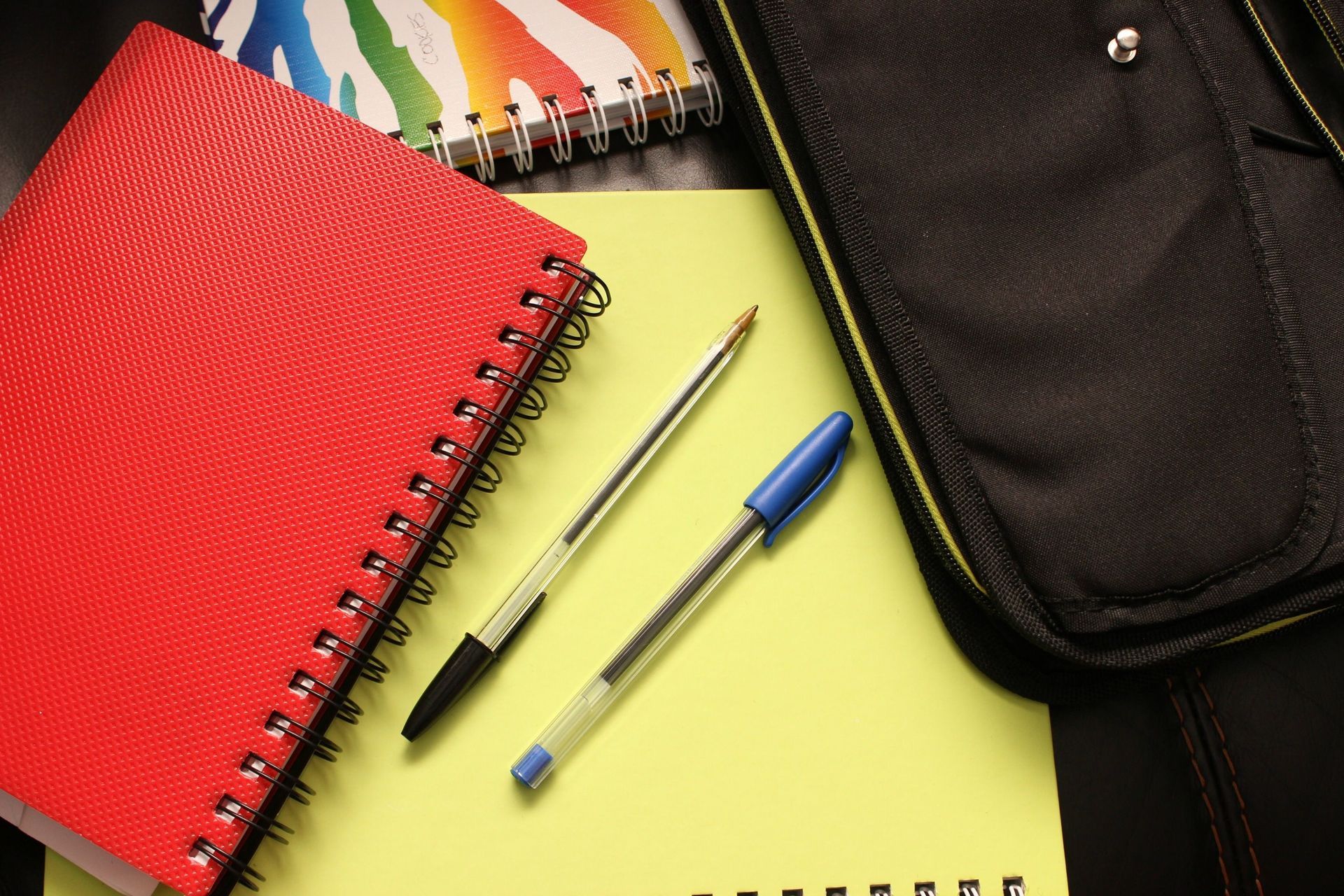 Un cuaderno de espiral rojo, bolígrafos y un estuche negro sobre un escritorio amarillo.