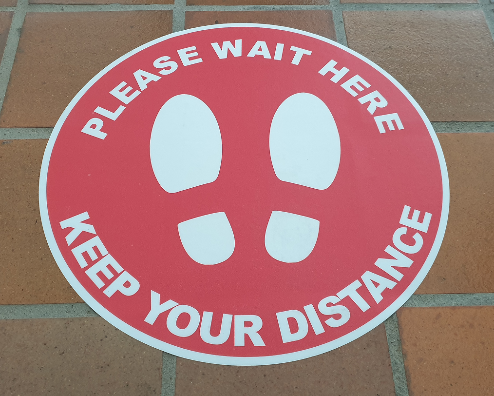 A red and white sign that says please wait here keep your distance
