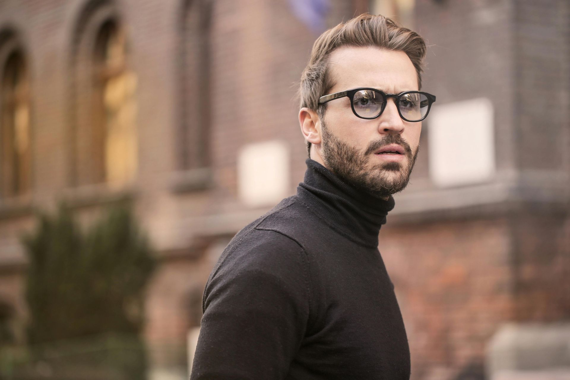 A man wearing glasses and a black turtleneck is standing in front of a building.