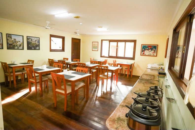Sala de jantar iluminada com mesas e cadeiras de madeira, quadros na parede e uma fileira de cafeteiras em um balcão lateral.
