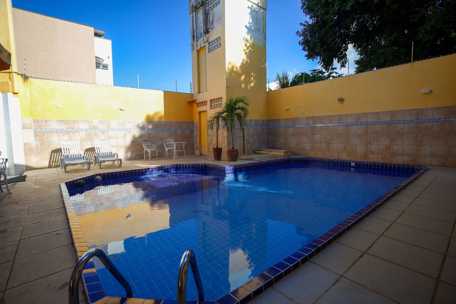 Pequeno pátio revestido com azulejos, piscina azul, paredes bege e uma palmeira sob a luz do sol.