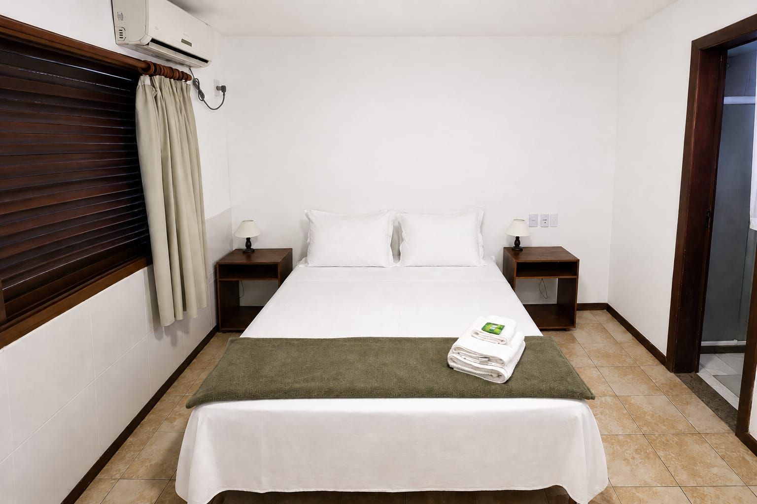 Quarto de hotel impecável com cama branca, manta verde, mesas de cabeceira e janela com cortinas.