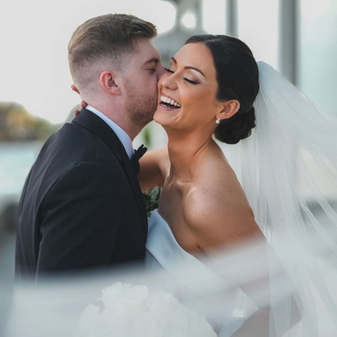 Groom kissing smiling bride on the cheek; veil blowing in wind. They are embraced outside on wedding day.