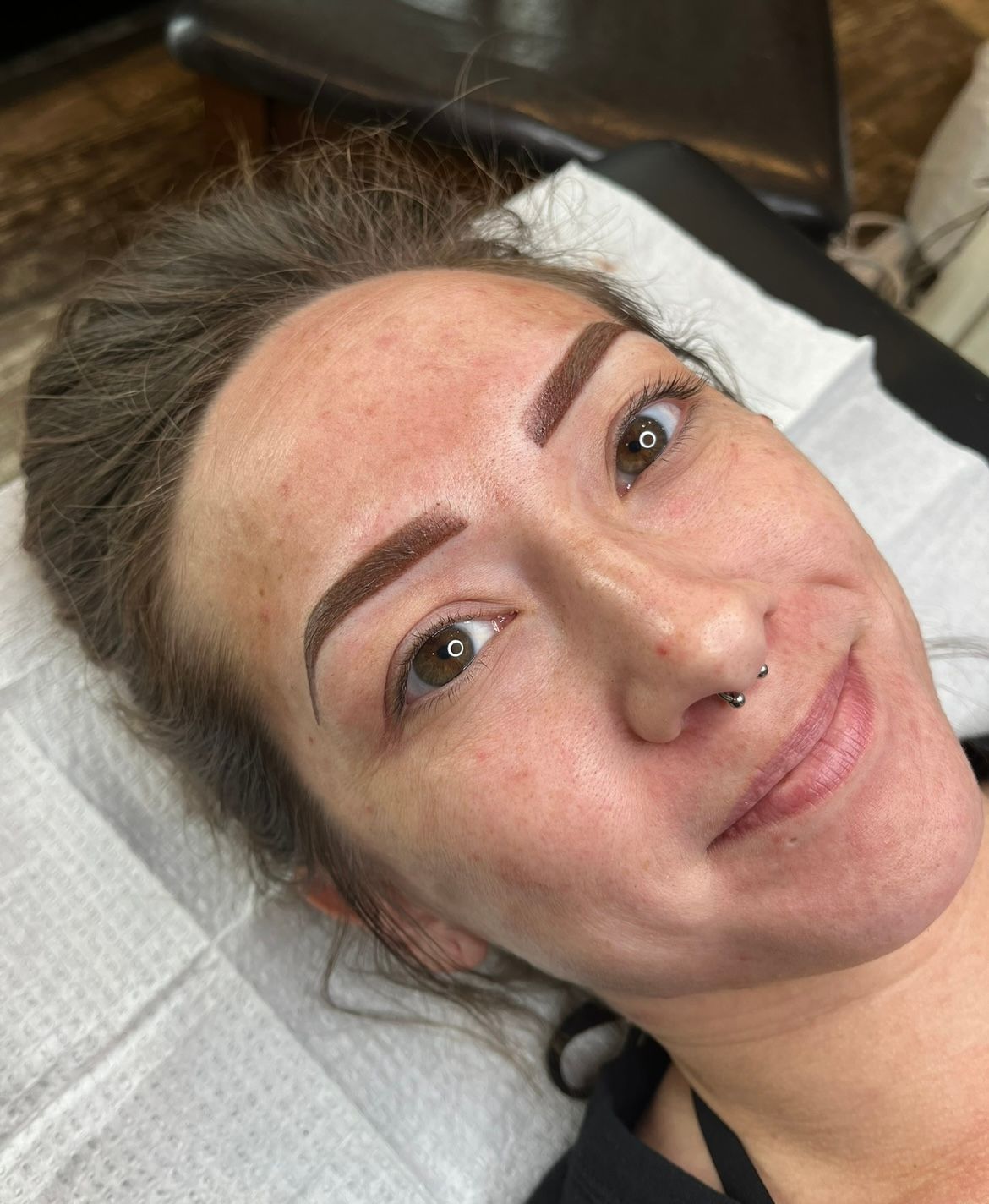 Woman with freshly microbladed eyebrows smiles while lying down on a white paper-covered table.