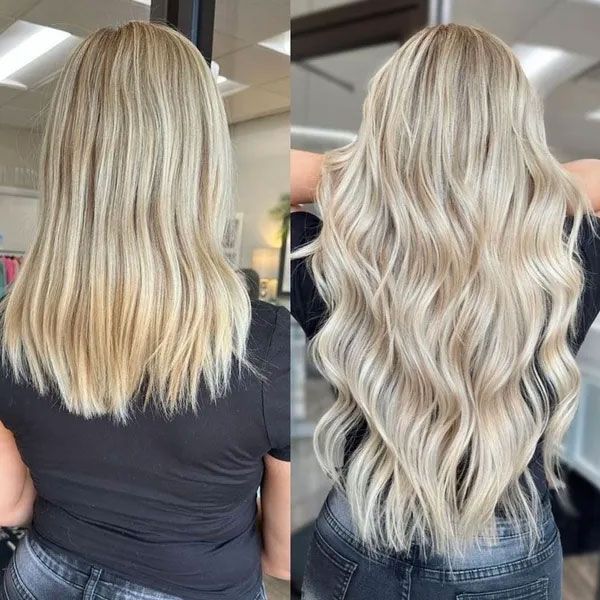 Long wavy hair with a dark root transition into blended blonde highlights, seen from behind in a salon setting.