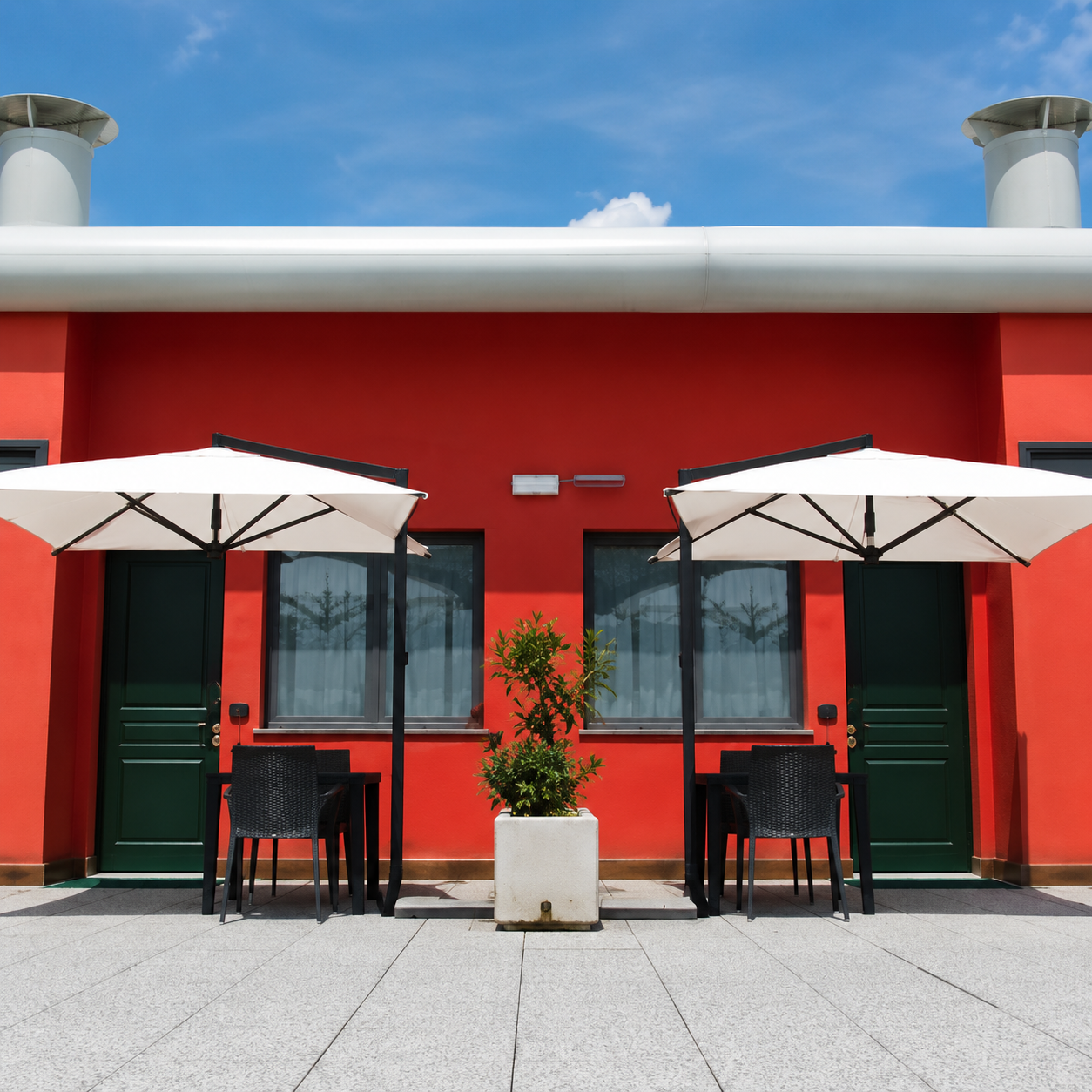 Facciata rossa del caffè con due ombrelloni bianchi e tavolini all'aperto su un patio soleggiato.