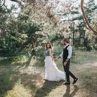 A bride and groom are walking through a forest holding hands.