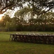 A row of tables and chairs in a field with string lights hanging from trees.