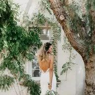 A woman in a wedding dress is standing next to a tree in front of a white building.