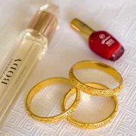 A bottle of perfume , a bottle of nail polish , and three gold bangles are on a table.