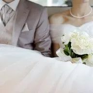 A bride and groom are sitting next to each other on a bed with a bouquet of flowers.