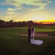 A bride and groom are standing on a golf course at sunset.