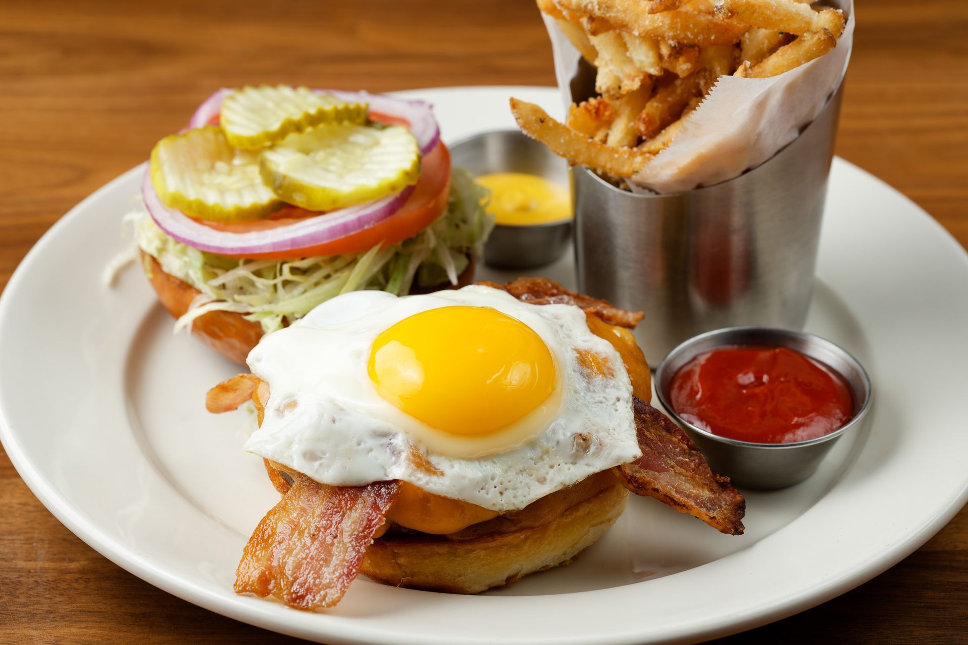 A hamburger with a fried egg , bacon , pickles and french fries on a plate.