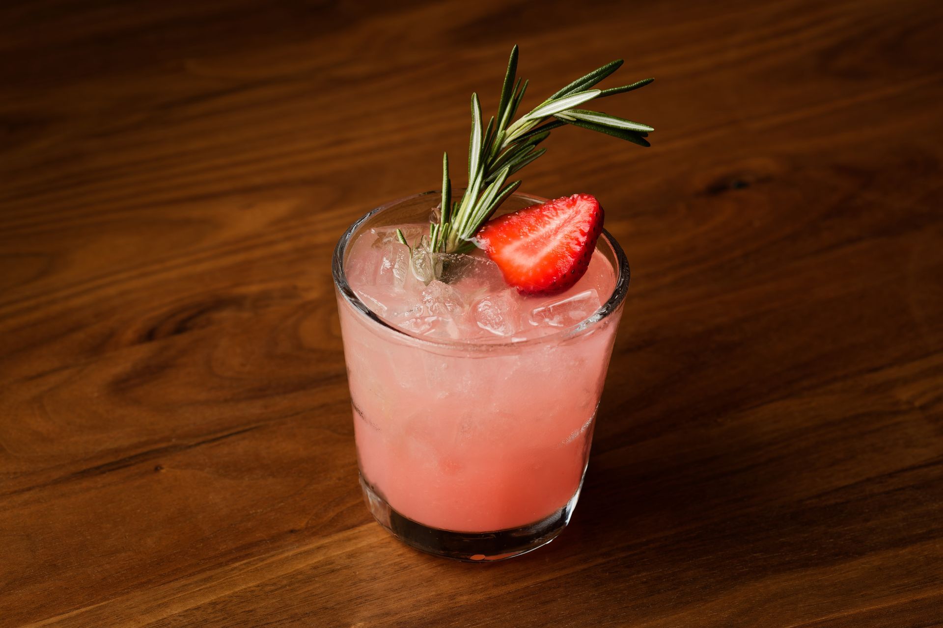 A glass of pink liquid with a strawberry and rosemary on a wooden table.