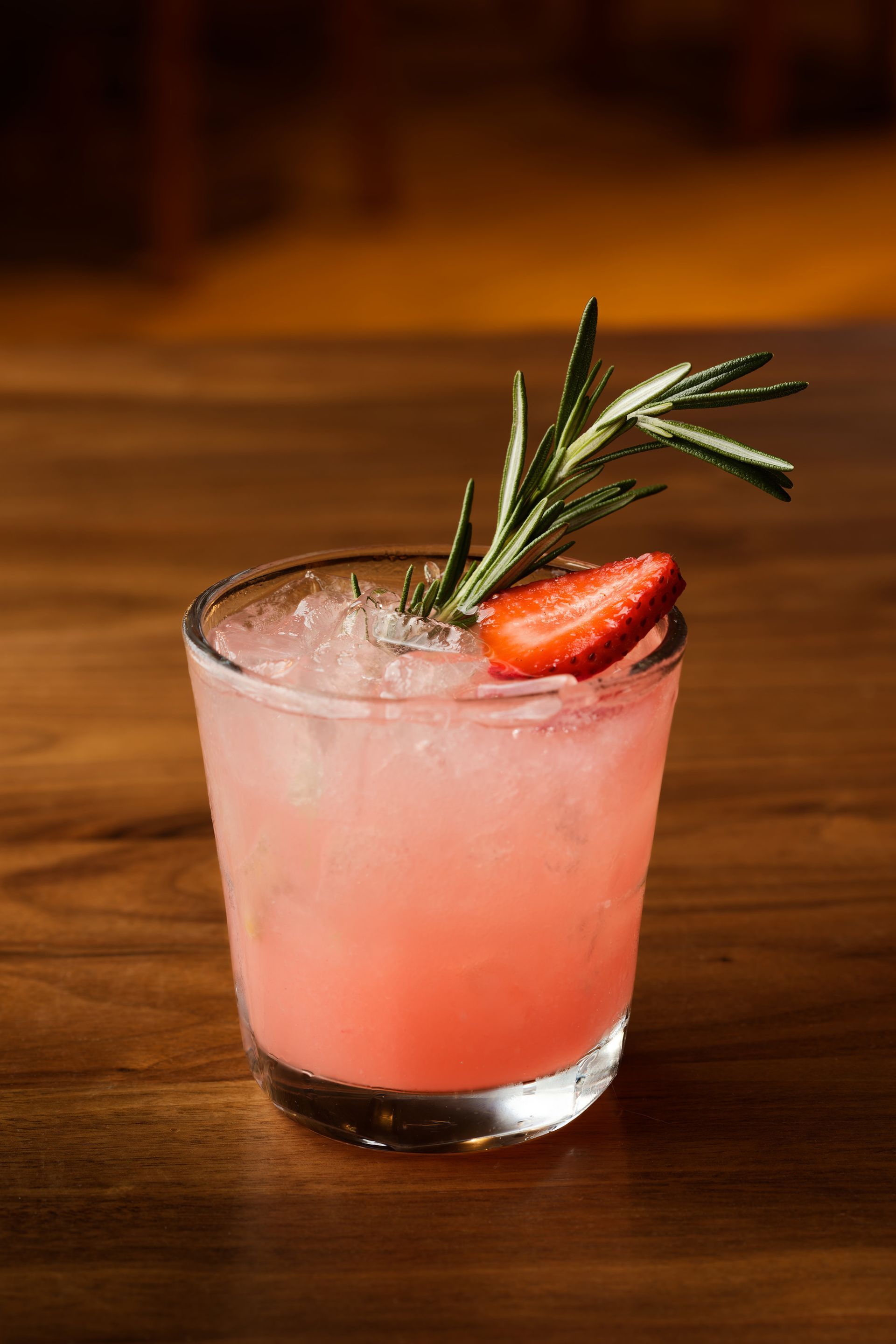 A glass of pink liquid with a strawberry and rosemary on a wooden table.